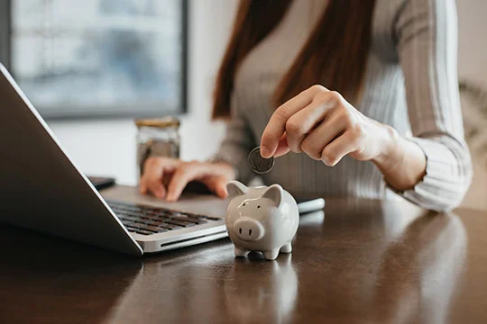 Mujer colocando una moneda en una alcancía frente a una laptop, simbolizando el ahorro gracias a una página web.
