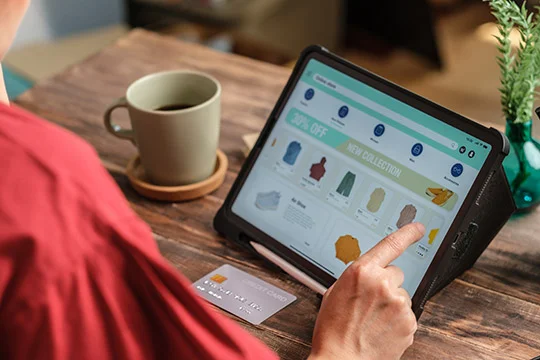 Persona navegando una tienda virtual en una tablet, eligiendo productos de moda con tarjeta a la mano.